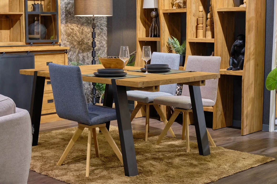 Une grande table de repas en bois et métal, dressée et entourée de chaises de repas, l'ensemble au sein d'un espace salle à manger décoré d'une lampe, d'une grande étagère en bois remplie de différents objets décoratifs. 