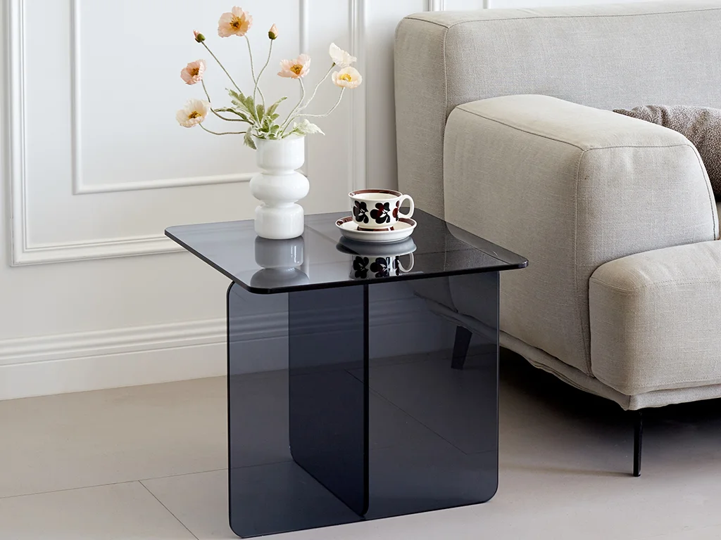 Une table en verre trempé bleue, élégante et raffinée, sur laquelle sont entreposés un petit vase blanc accueillant des fleurs orange, et à côté, une petite tasse de thé.