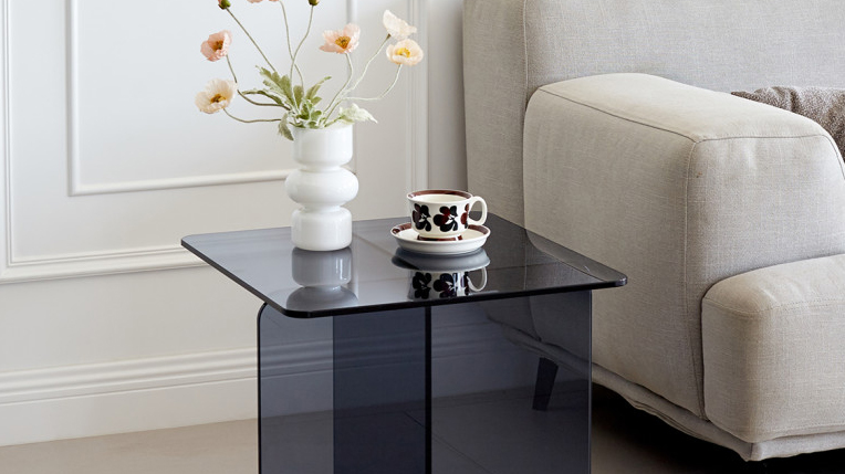Photo d'une table d'appoint en verre trempé d'un bleu marine élégant. La table est installée à côté d'un canapé de couleur gris clair, et elle accueille un petit vase avec des fleurs oranges ainsi qu'une tasse à thé dans sa coupelle.